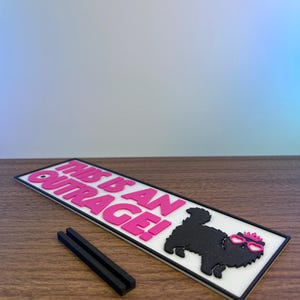 Puede incluir: Un letrero rectangular blanco con el texto "THIS IS AN OUTRAGE!" en rosa. Un perro negro con gafas rosas y un lazo rosa est&aacute; en el lado derecho. Dos piezas rectangulares negras est&aacute;n frente al letrero.