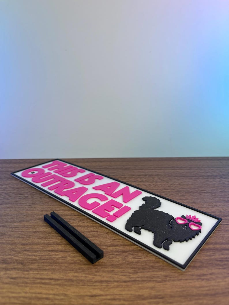 Puede incluir: Un letrero rectangular blanco con el texto "THIS IS AN OUTRAGE!" en rosa. Un perro negro con gafas rosas y un lazo rosa est&aacute; en el lado derecho. Dos piezas rectangulares negras est&aacute;n frente al letrero.