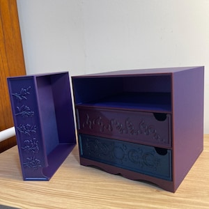 May include: A purple two-drawer storage box with a matching side compartment. The drawers have decorative floral and celestial designs. The box is made of a solid material and is suitable for small items.