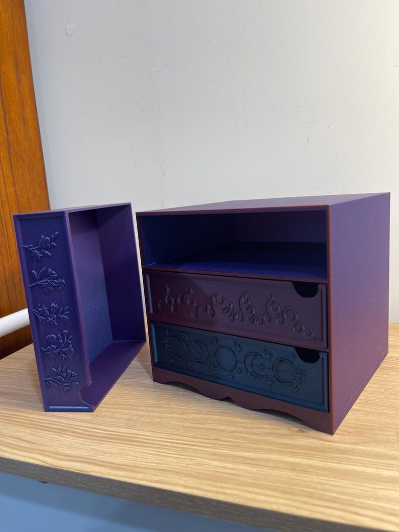 May include: A purple two-drawer storage box with a matching side compartment. The drawers have decorative floral and celestial designs. The box is made of a solid material and is suitable for small items.