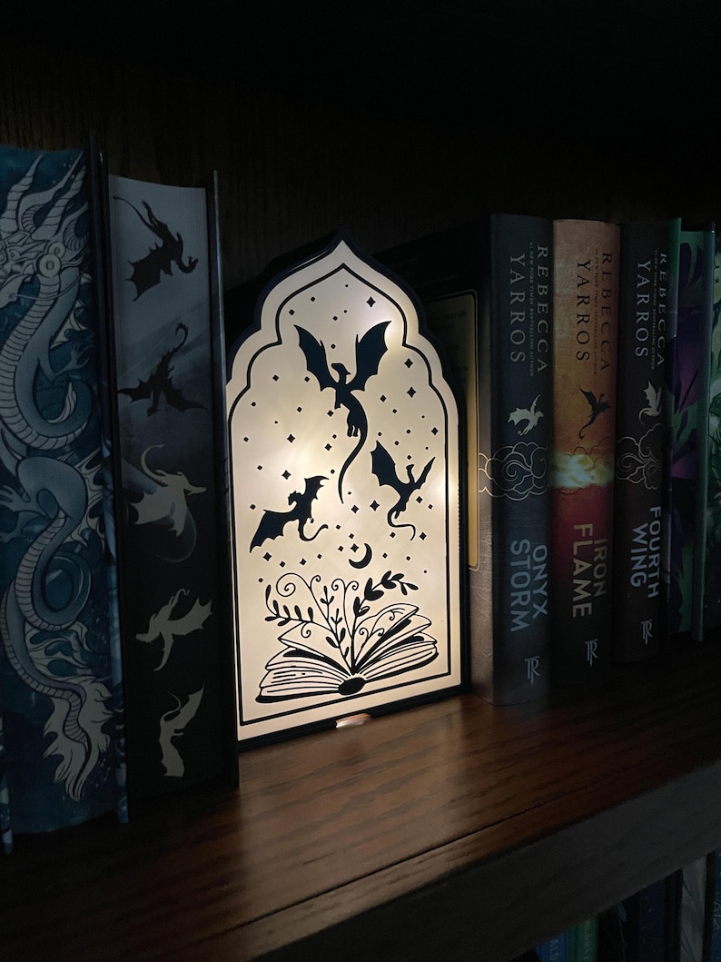 Op de afbeelding: Een decoratieve, verlichte boekensteun met een zwart silhouet van draken, sterren en een open boek met bladeren. De boekensteun staat op een houten plank met boeken, wat een fantasiethema cre&euml;ert.