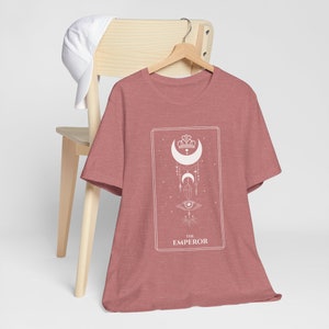 May include: A heather red t-shirt featuring a white graphic of a tarot card, "The Emperor", with a crescent moon, crown, and eye symbol.