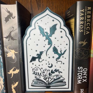 Op de afbeelding: Een witte en blauwgroene boekensteun met drakenmotief en een decoratieve boog. De boekensteun heeft drie draken, sterren en een open boek met bladeren. Het staat tussen boeken op een houten plank.