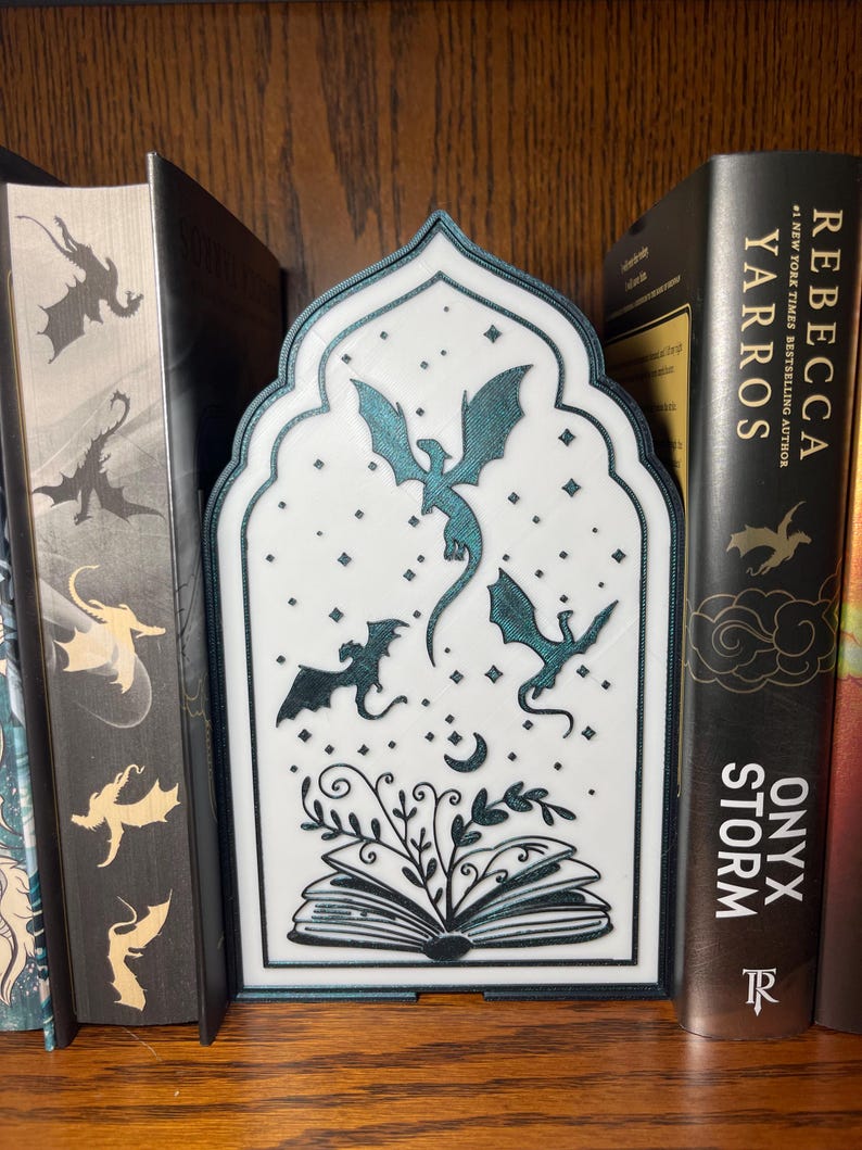 Op de afbeelding: Een witte en blauwgroene boekensteun met drakenmotief en een decoratieve boog. De boekensteun heeft drie draken, sterren en een open boek met bladeren. Het staat tussen boeken op een houten plank.