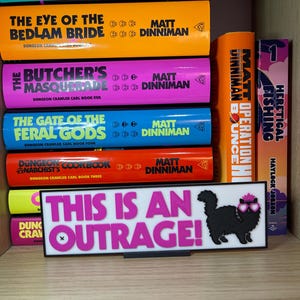 Puede incluir: Una estanter&iacute;a muestra libros con lomos coloridos, con t&iacute;tulos como "This Inevitable Ruin" y "The Eye of the Bedlam Bride". Un letrero con el texto "THIS IS AN OUTRAGE!" y un gato con gafas de sol tambi&eacute;n es visible.