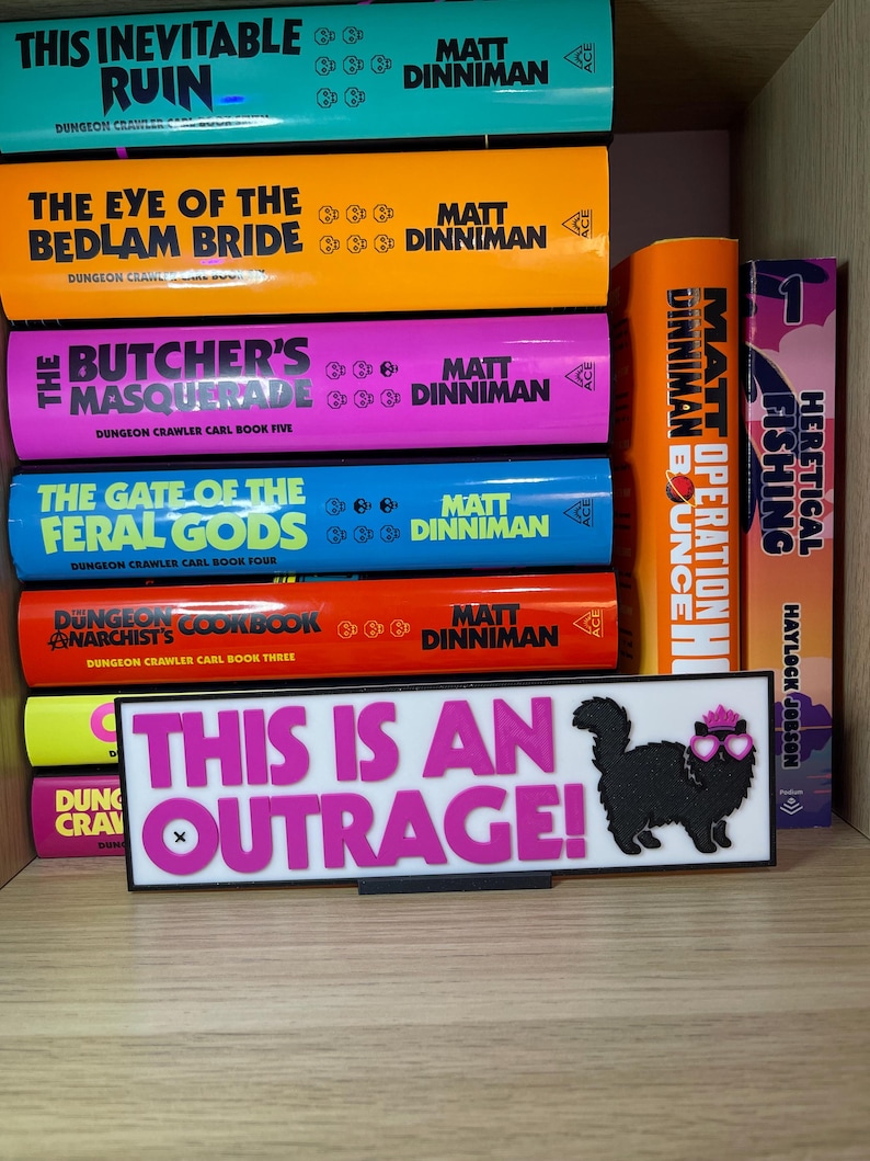 Puede incluir: Una estanter&iacute;a muestra libros con lomos coloridos, con t&iacute;tulos como "This Inevitable Ruin" y "The Eye of the Bedlam Bride". Un letrero con el texto "THIS IS AN OUTRAGE!" y un gato con gafas de sol tambi&eacute;n es visible.