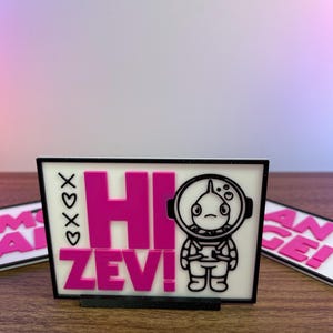 Puede incluir: Un letrero rectangular con el texto "HI ZEV!" en rosa brillante, acompa&ntilde;ado de una ilustraci&oacute;n de un astronauta de dibujos animados. El letrero tiene un borde negro y est&aacute; sobre una superficie de madera. Tambi&eacute;n son visibles las letras "xoxo".