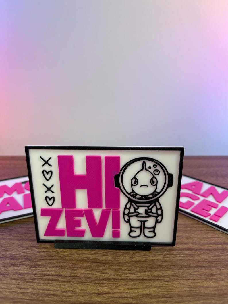 Puede incluir: Un letrero rectangular con el texto "HI ZEV!" en rosa brillante, acompa&ntilde;ado de una ilustraci&oacute;n de un astronauta de dibujos animados. El letrero tiene un borde negro y est&aacute; sobre una superficie de madera. Tambi&eacute;n son visibles las letras "xoxo".