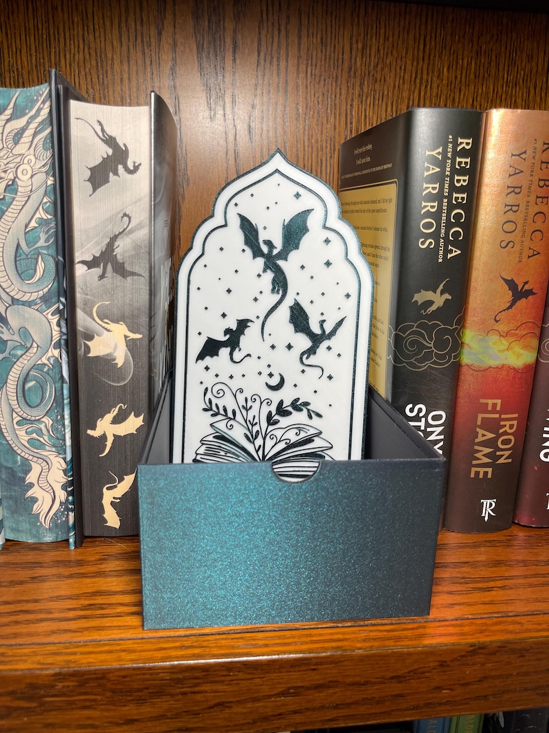 Op de afbeelding: Een fantasy-thema bladwijzer met een drakenontwerp. De bladwijzer is wit met zwarte draken silhouetten en een zwarte doos. De bladwijzer wordt in een doos op een houten plank met boeken weergegeven.