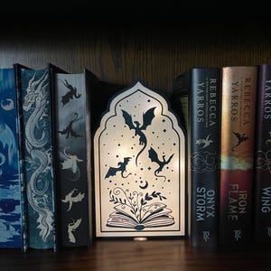 Op de afbeelding: Een decoratieve houten lamp met een draakthema, met een silhouet van draken, een open boek en een maan. De lamp staat op een boekenplank met boeken, waaronder "Onyx Storm" en "Iron Flame".