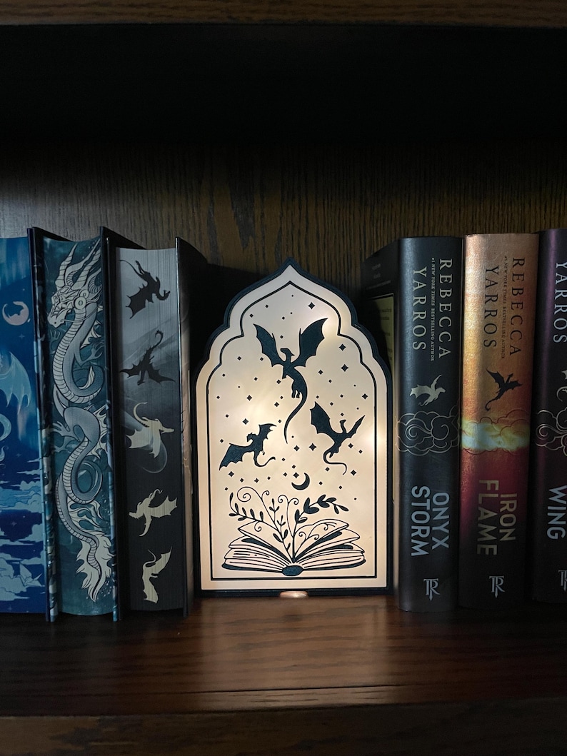 Op de afbeelding: Een decoratieve houten lamp met een draakthema, met een silhouet van draken, een open boek en een maan. De lamp staat op een boekenplank met boeken, waaronder "Onyx Storm" en "Iron Flame".