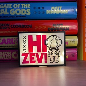 Puede incluir: Una luz LED rectangular con el texto "HI ZEV!" en rosa brillante y una ilustraci&oacute;n de un astronauta de dibujos animados. La luz, enmarcada en negro, est&aacute; frente a una pila de libros coloridos.