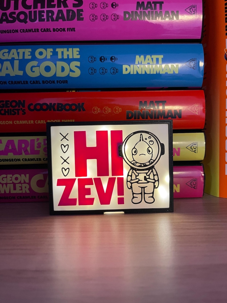 Puede incluir: Una luz LED rectangular con el texto "HI ZEV!" en rosa brillante y una ilustraci&oacute;n de un astronauta de dibujos animados. La luz, enmarcada en negro, est&aacute; frente a una pila de libros coloridos.