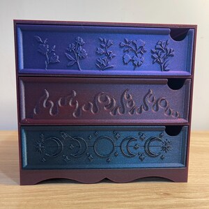 May include: A three-drawer storage chest with a gradient color scheme. The top drawer features floral designs, the middle drawer has flame motifs, and the bottom drawer displays crescent moon and star patterns. The chest is primarily purple, with blue and red accents.