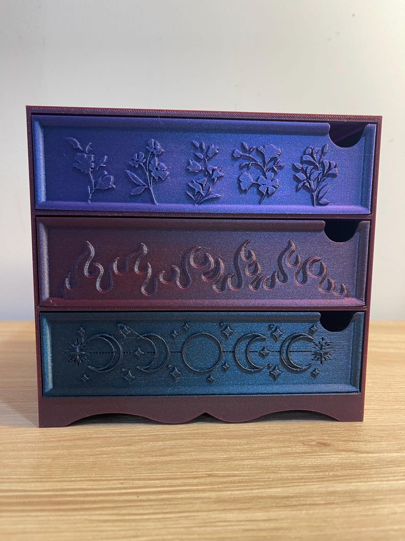 May include: A three-drawer storage chest with a gradient color scheme. The top drawer features floral designs, the middle drawer has flame motifs, and the bottom drawer displays crescent moon and star patterns. The chest is primarily purple, with blue and red accents.