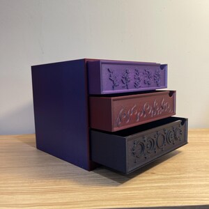 May include: A three-drawer storage box in shades of purple, burgundy, and dark gray. Each drawer is decorated with a different embossed design: botanical, swirling lines, and celestial motifs. The box sits on a wooden surface.