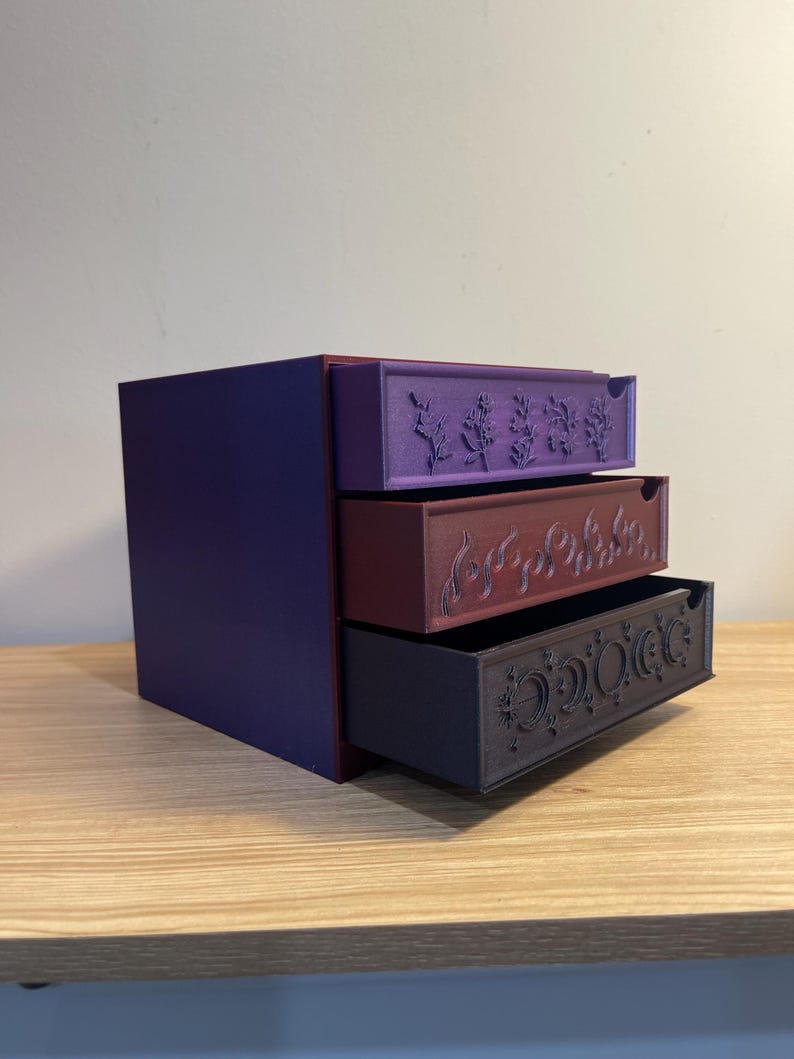 May include: A three-drawer storage box in shades of purple, burgundy, and dark gray. Each drawer is decorated with a different embossed design: botanical, swirling lines, and celestial motifs. The box sits on a wooden surface.