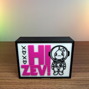 Puede incluir: Una caja negra con un panel frontal blanco. El panel muestra el texto rosa "HI ZEV!", un astronauta de dibujos animados y corazones con las letras "XOXO".