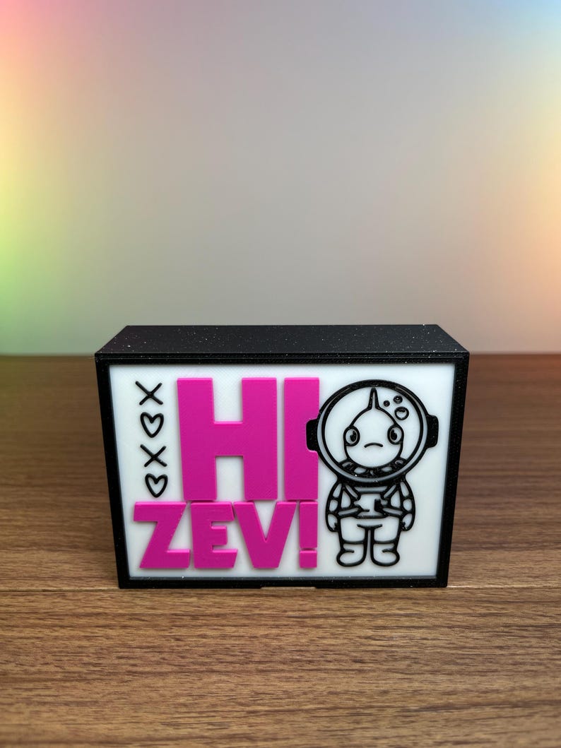 Puede incluir: Una caja negra con un panel frontal blanco. El panel muestra el texto rosa "HI ZEV!", un astronauta de dibujos animados y corazones con las letras "XOXO".