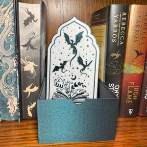 Op de afbeelding: Een teal en zwarte boekenleggerhouder met een bijpassende boekenlegger met een drakenontwerp. De boekenlegger is wit met zwarte drakensilhouetten en bloemmotieven. Op een houten plank met boeken.