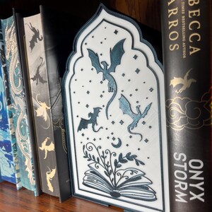 Op de afbeelding: Een witte en blauwgroene boekensteun met een draakthema en een decoratief boogontwerp. De boekensteun heeft een gedetailleerde illustratie van draken, sterren en een open boek met bladeren. Het staat op een houten plank met verschillende boeken.