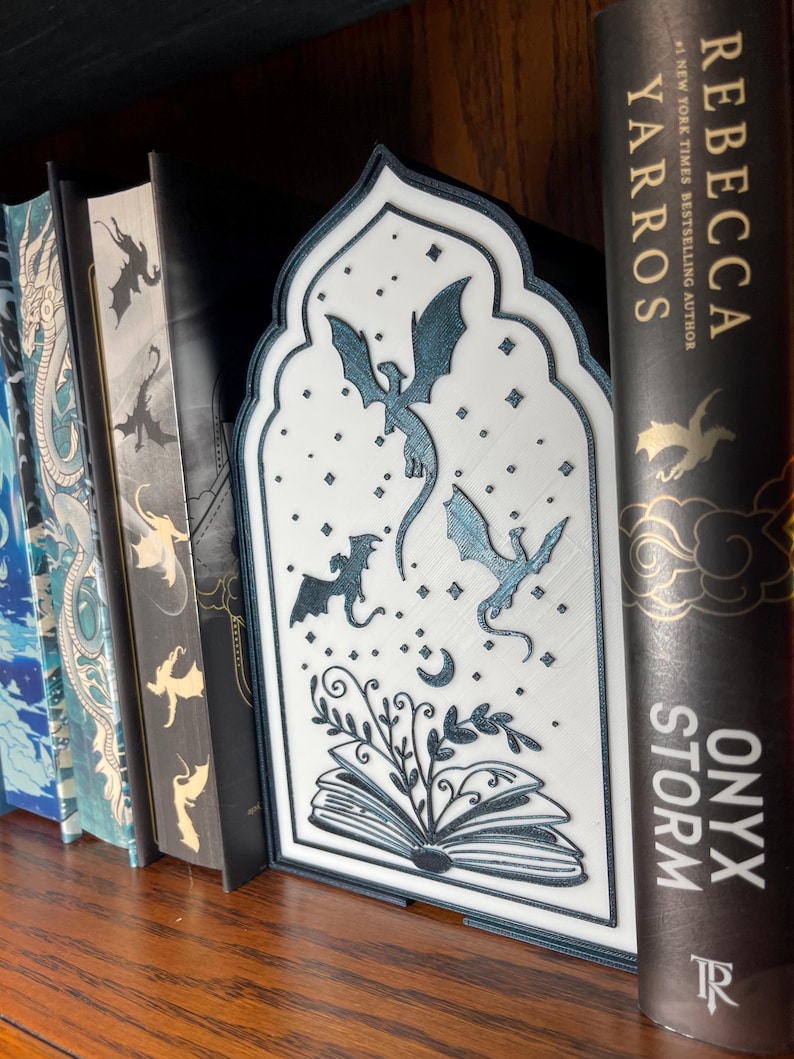 Op de afbeelding: Een witte en blauwgroene boekensteun met een draakthema en een decoratief boogontwerp. De boekensteun heeft een gedetailleerde illustratie van draken, sterren en een open boek met bladeren. Het staat op een houten plank met verschillende boeken.