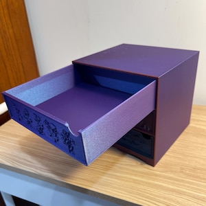 May include: A small, purple wooden box with a drawer partially open. The drawer's interior has a shimmering, light purple finish. The box has a dark purple exterior and a decorative design on the drawer's front.