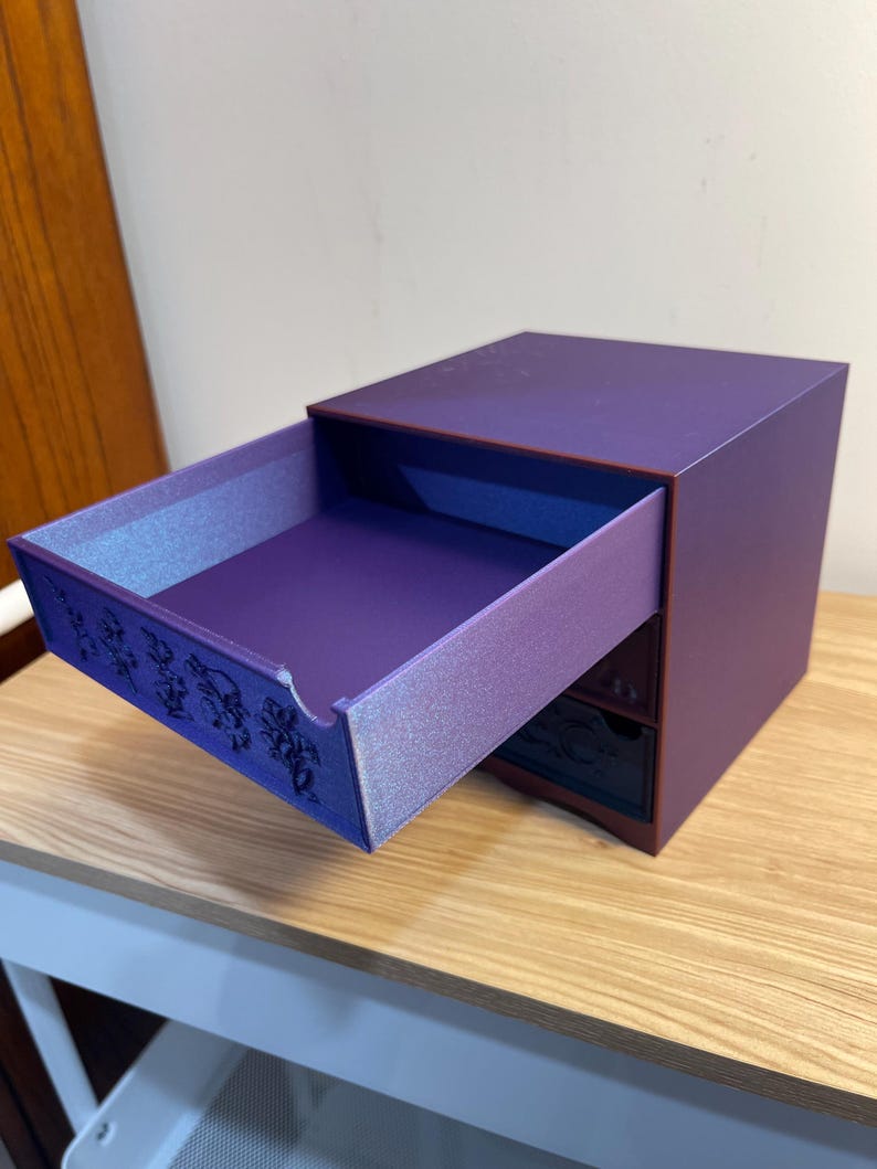 May include: A small, purple wooden box with a drawer partially open. The drawer's interior has a shimmering, light purple finish. The box has a dark purple exterior and a decorative design on the drawer's front.