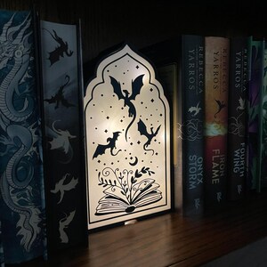 Op de afbeelding: Een decoratieve zwart-witte LED-lamp met een drakenontwerp, geplaatst op een houten plank tussen boeken. De lamp heeft silhouetten van draken, een open boek en een maan. De boeken hebben draken- en fantasiethema's.