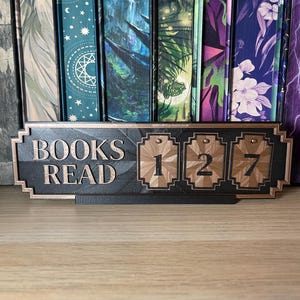 May include: A decorative sign with the words "BOOKS READ" and the numbers "127" displayed. The sign is black with copper-colored accents and is placed in front of a row of colorful books.