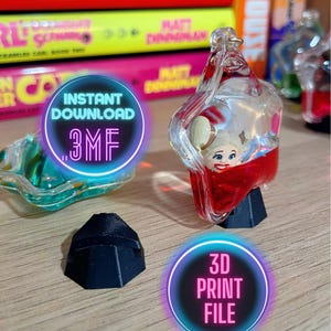 May include: A clear, decorative bottle stopper with a cartoon character inside, resting on a black base. The stopper has red liquid at the bottom. The image also includes a separate black base and text that says "Instant Download .3MF" and "3D Print File".