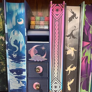 May include: A dark blue storage unit with two drawers and a decorative moon and star design. The unit is flanked by colorful books with dragon and celestial illustrations. The drawers have pink accents and gold knobs. A set of colorful keycaps sits on top.