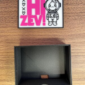 Puede incluir: Un parche rectangular con el texto "HI ZEV!" en rosa, y una ilustraci&oacute;n de un astronauta. El parche est&aacute; sobre un fondo blanco con un borde negro. Debajo hay una caja rectangular negra con un peque&ntilde;o recorte.