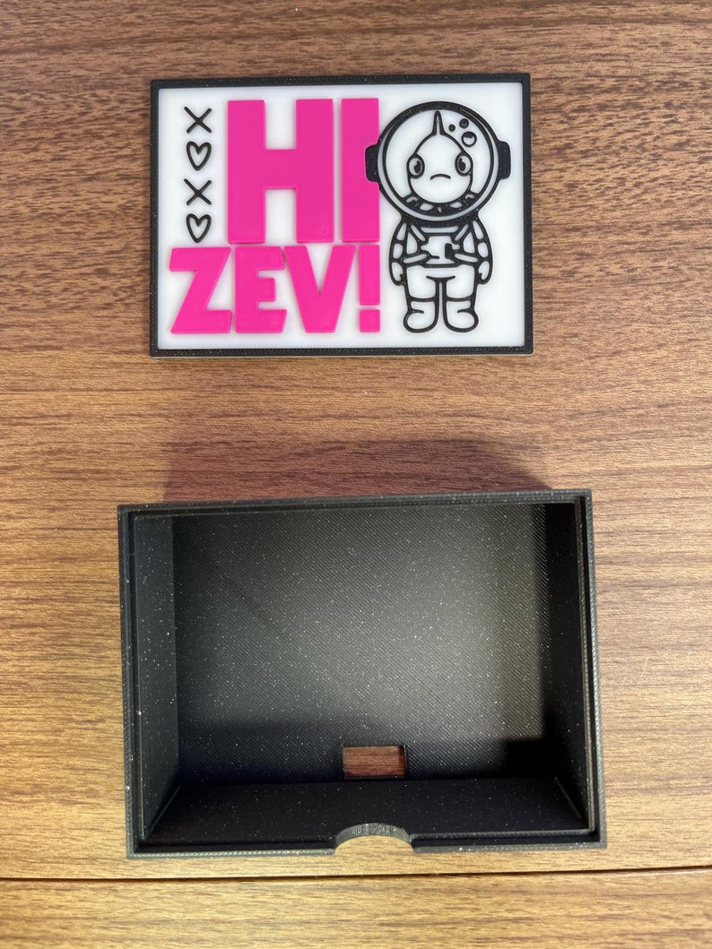 Puede incluir: Un parche rectangular con el texto "HI ZEV!" en rosa, y una ilustraci&oacute;n de un astronauta. El parche est&aacute; sobre un fondo blanco con un borde negro. Debajo hay una caja rectangular negra con un peque&ntilde;o recorte.