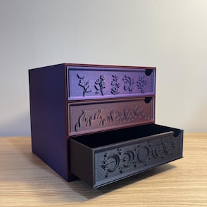 May include: A three-drawer storage box with a purple exterior. Each drawer is a different color: purple, maroon, and black. The drawers feature embossed designs of plants, flames, and phases of the moon. The box is on a wooden surface.