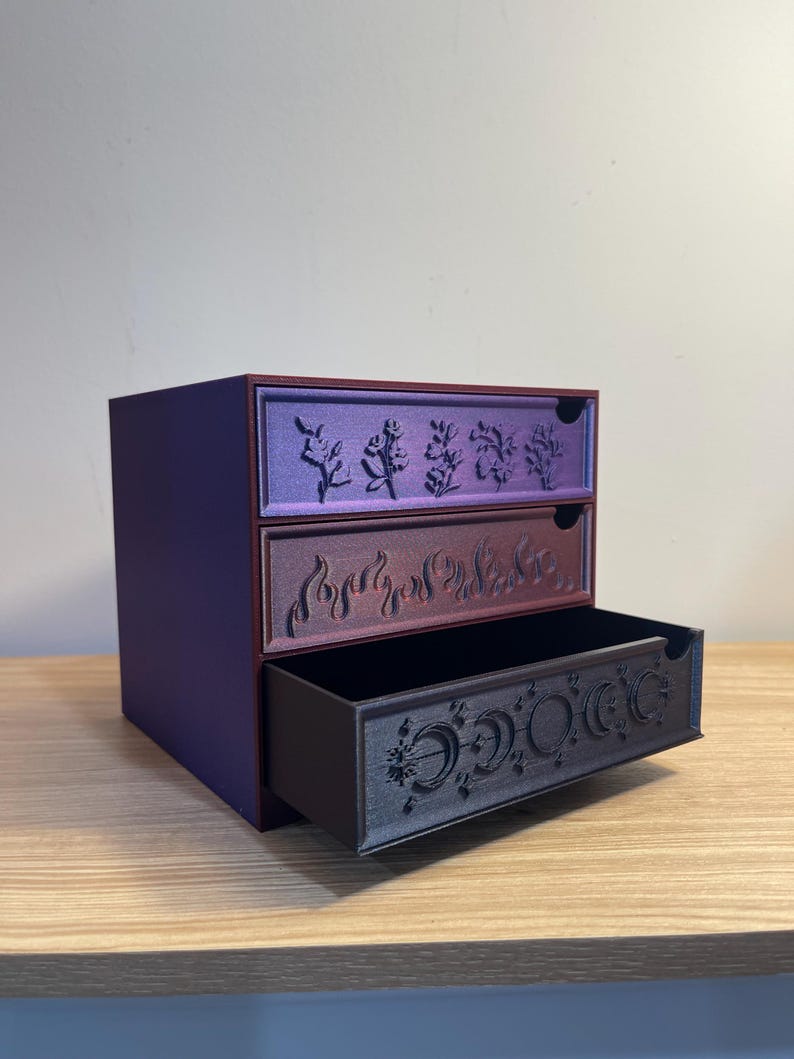 May include: A three-drawer storage box with a purple exterior. Each drawer is a different color: purple, maroon, and black. The drawers feature embossed designs of plants, flames, and phases of the moon. The box is on a wooden surface.