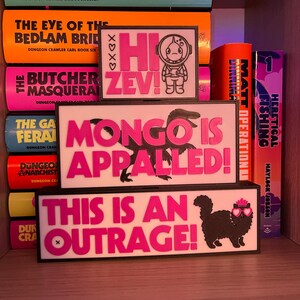 Puede incluir: Una estanter&iacute;a exhibe libros y tres letreros rectangulares con bordes negros. Los letreros presentan texto e ilustraciones en rosa. Uno dice "Hi Zev!" con un astronauta, otro "Mongo is Appalled!", y el &uacute;ltimo "This is an Outrage!" con un gato.