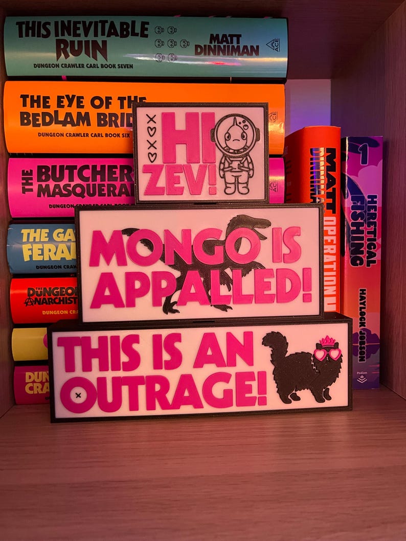Puede incluir: Una estanter&iacute;a exhibe libros y tres letreros rectangulares con bordes negros. Los letreros presentan texto e ilustraciones en rosa. Uno dice "Hi Zev!" con un astronauta, otro "Mongo is Appalled!", y el &uacute;ltimo "This is an Outrage!" con un gato.