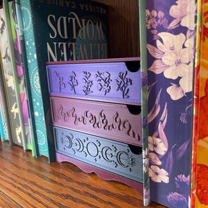 May include: Three-drawer storage unit in shades of purple, with decorative cut-out designs. The top drawer features floral patterns, the middle drawer has flames, and the bottom drawer displays crescent moons and stars. The unit sits on a wooden shelf next to a row of books.