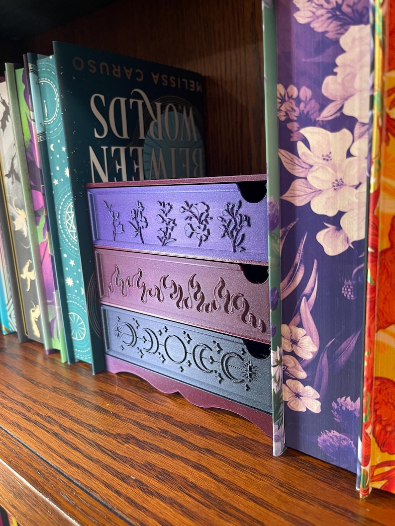 May include: Three-drawer storage unit in shades of purple, with decorative cut-out designs. The top drawer features floral patterns, the middle drawer has flames, and the bottom drawer displays crescent moons and stars. The unit sits on a wooden shelf next to a row of books.