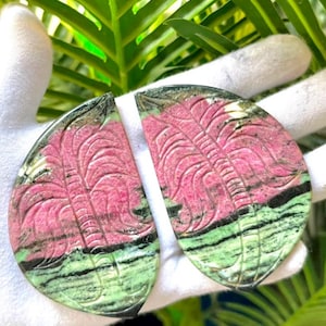 Puede incluir: Dos cabujones de piedra pulida en forma de semicírculo con un diseño de hoja de palmera tallada. Las piedras muestran una paleta de colores vibrantes con bandas horizontales en rosa oscuro, verde claro y negro, con texturas minerales.