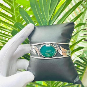 Könnte beinhalten: Ein silbernes Armband mit einem zentralen ovalen grünen Stein in einer gezackten Fassung. Das Armband besteht aus zwei parallelen Bändern und zwei silbernen Platten auf beiden Seiten des Steins, die mit arabischer Schrift graviert sind.