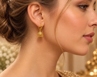 Könnte beinhalten: Ein goldener Creolen-Ohrring mit einem kleinen, glockenförmigen Jhumka-Anhänger. Der Anhänger weist ein filigranes Muster mit winzigen Aussparungen und einem gewellten unteren Rand auf und hängt an einem schlichten, polierten Goldring.