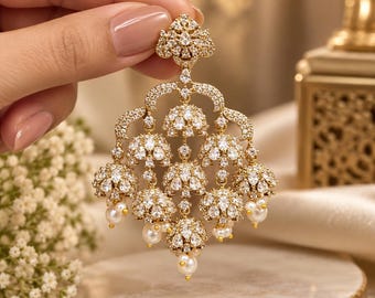 May include: A gold-toned chandelier earring featuring a triangular top stud and a multi-tiered structure adorned with numerous clear, faceted crystals. The design includes several bell-shaped elements accented with small, round white pearls.