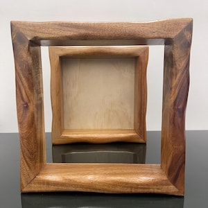 May include: A square wooden frame with a natural, wavy edge finish and a rich brown wood grain pattern. The frame is positioned in front of a smaller, square wooden box with a light-colored interior base, creating a nested visual effect.