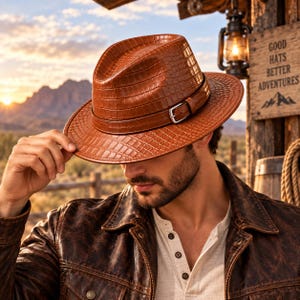May include: A brown fedora hat with a textured crocodile skin pattern, featuring a matching brown hat band with a silver-toned metal buckle. The hat has a structured crown and a wide brim, showcasing a glossy finish and detailed embossed texture.