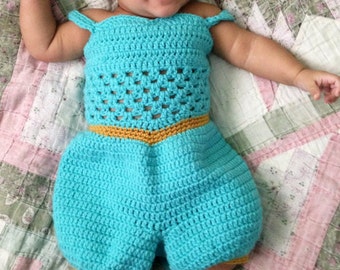 May include: A baby wearing a blue crocheted jumpsuit with a gold trim and a blue headband with a jewel. The baby is lying on a pink and green patterned blanket.