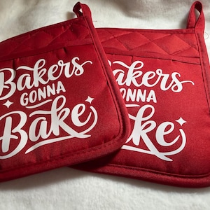 May include: Two square red oven mitts featuring a quilted texture and a hanging loop at the top corner. Each mitt displays the white printed text Bakers GONNA BAKE in a stylized script font, accented with small four-pointed star icons.