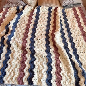 May include: A crocheted blanket featuring a repeating ripple pattern in alternating vertical stripes of cream, dusty rose, and slate blue yarn. The textile displays a consistent wavy texture throughout, showcasing the intricate interlocking stitches.