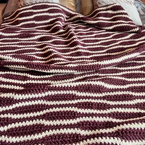 Könnte beinhalten: Eine handgehäkelte Decke mit einem sich wiederholenden Muster aus horizontalen Streifen in tiefem Burgunderrot und Cremeweiß. Das Textil weist eine dicke, strukturierte Maschenkonstruktion mit einem weichen, gefalteten Fall auf.